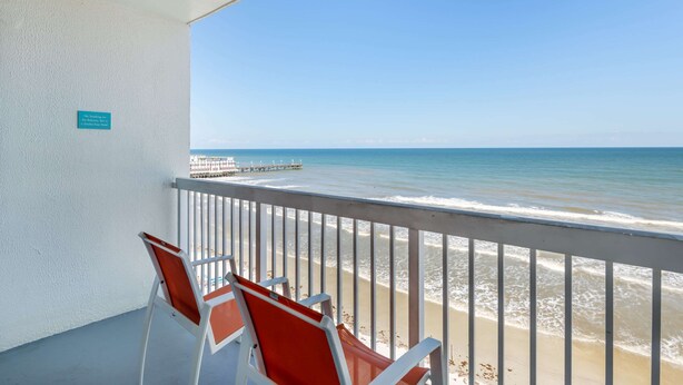 Ein Balkon mit Blick auf den Strand und den Pier. Zwei Stühle und ein Schild an der Wand sind zu sehen.