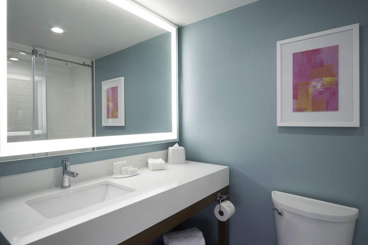 A bathroom with a white sink, mirror, and toilet. There is a framed artwork on the wall and a roll of toilet paper hanging on the side.
