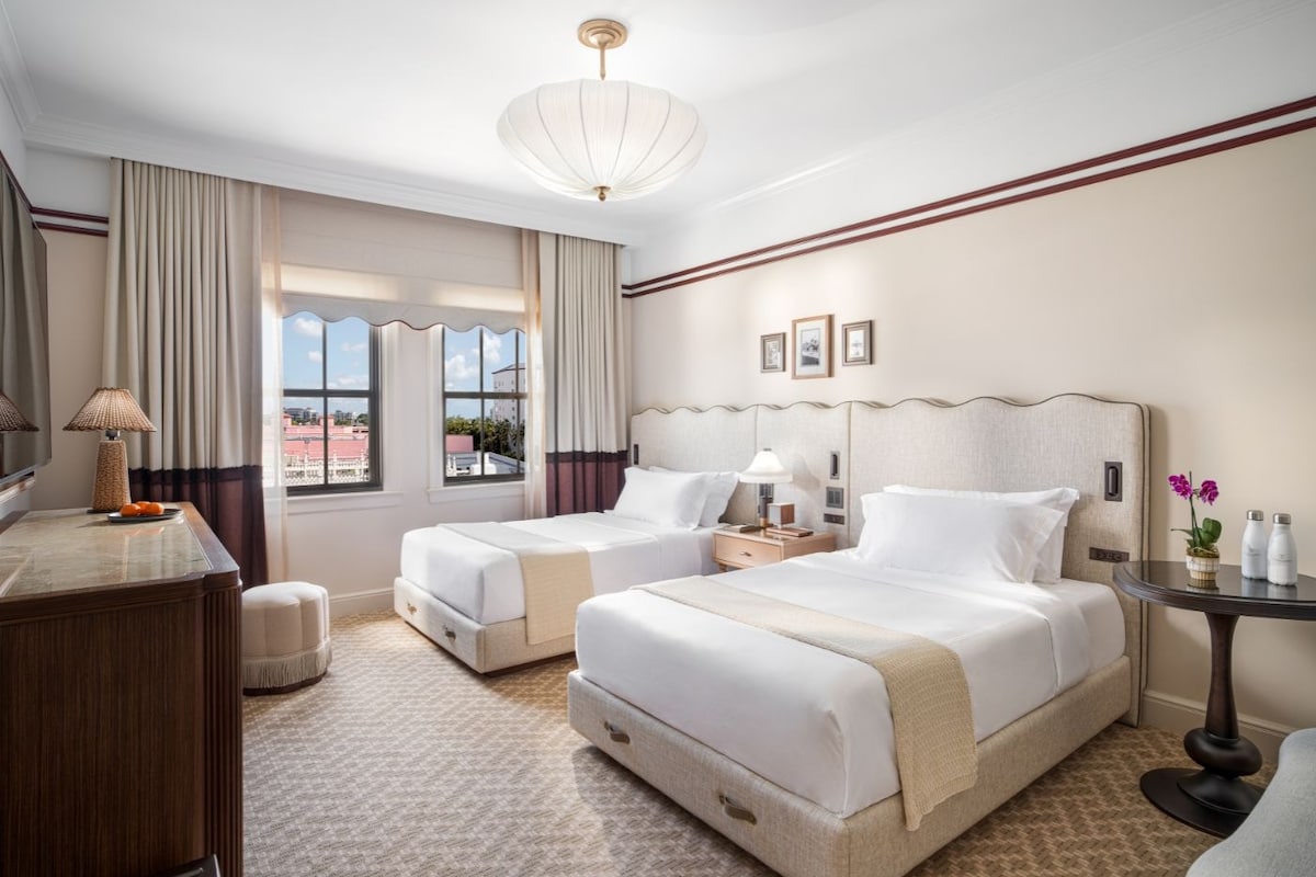 Habitación de hotel con dos camas, un escritorio, una silla, una mesita y vistas a la ciudad.
