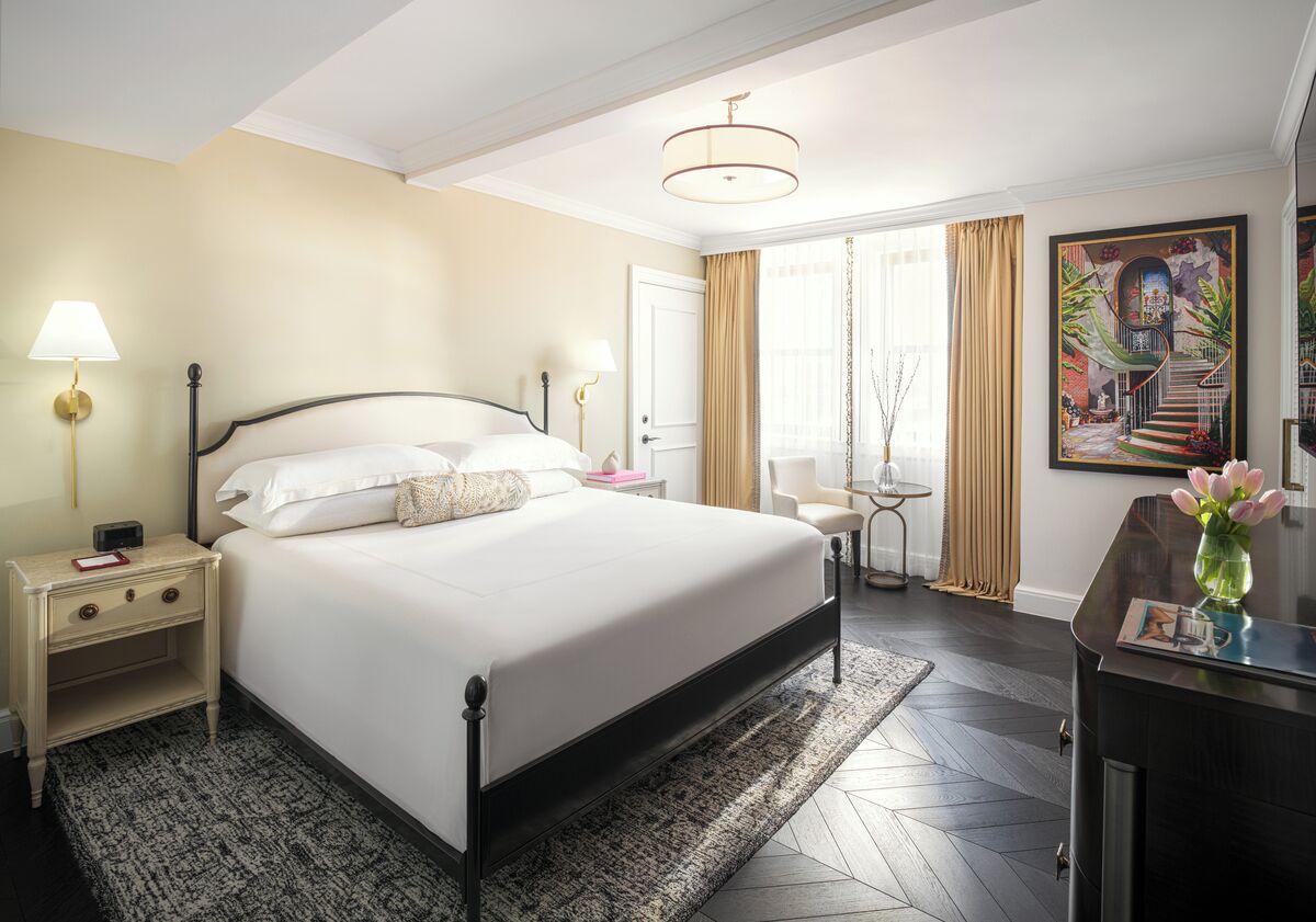 A hotel room with a large bed, bedside tables, a desk, a chair, a lamp, a painting, and a view of the outside.