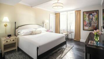 A hotel room with a large bed, bedside tables, a desk, a chair, a lamp, a painting, and a view of the outside.