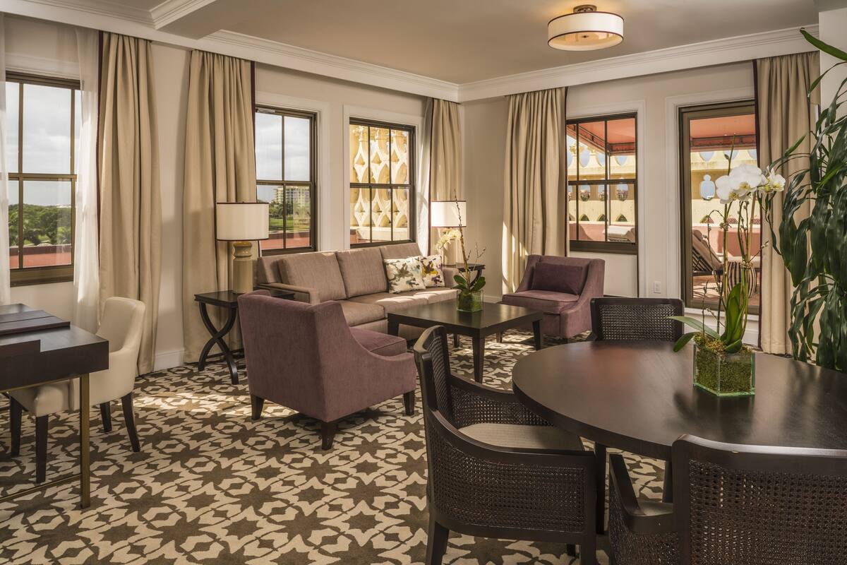 A hotel room with a sofa, chairs, a dining table, and a view of the cityscape through large windows.