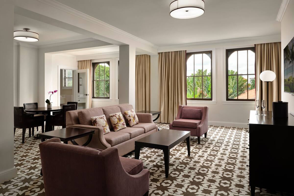 A modern hotel room with a sofa, armchairs, a dining table, and a TV.