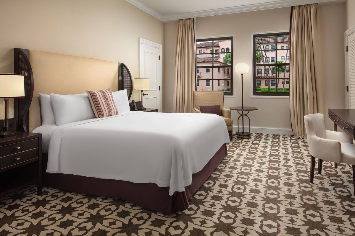 A hotel room with a large bed, a desk, a chair, a lamp, and a view of buildings through the window.
