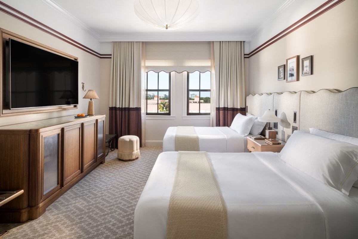 Habitación de hotel con un televisor de pantalla plana grande, dos camas, un armario de madera, un pequeño reposapiés y vistas al exterior a través de las ventanas.