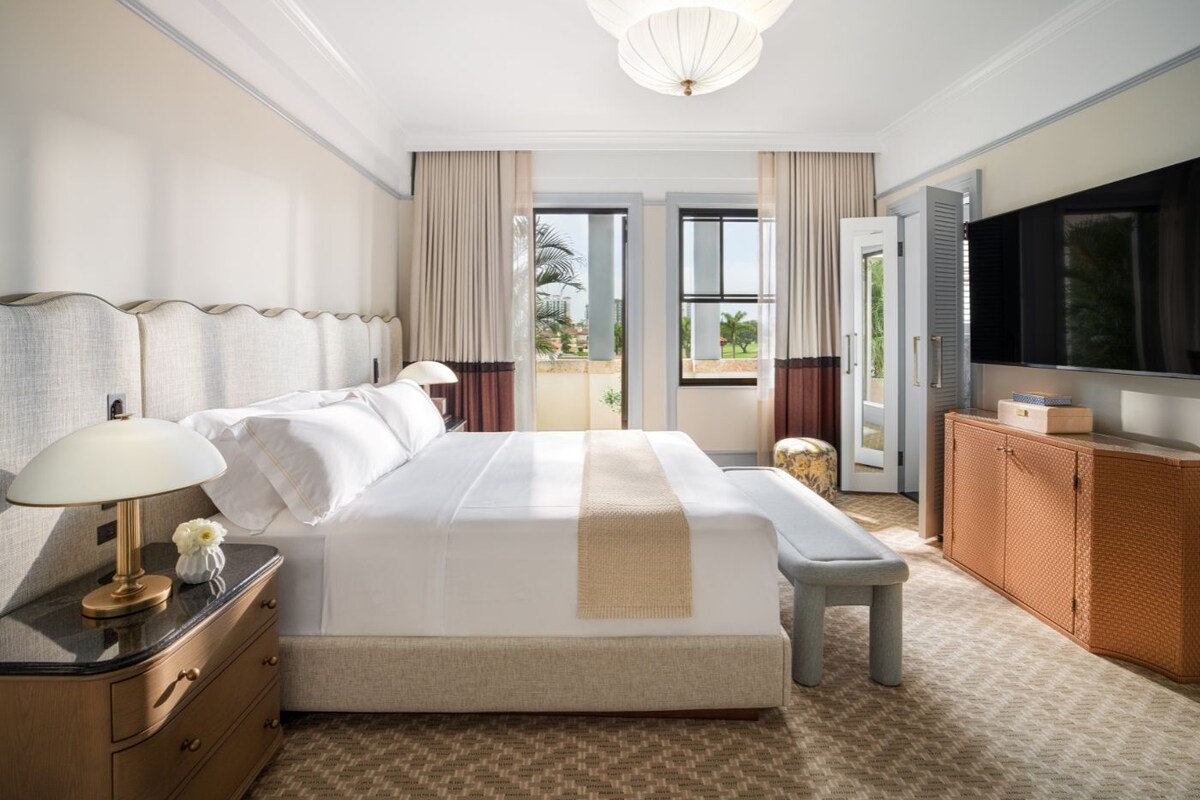 Habitación de hotel con una cama grande, televisión, escritorio con lámpara y vista al exterior desde la puerta.