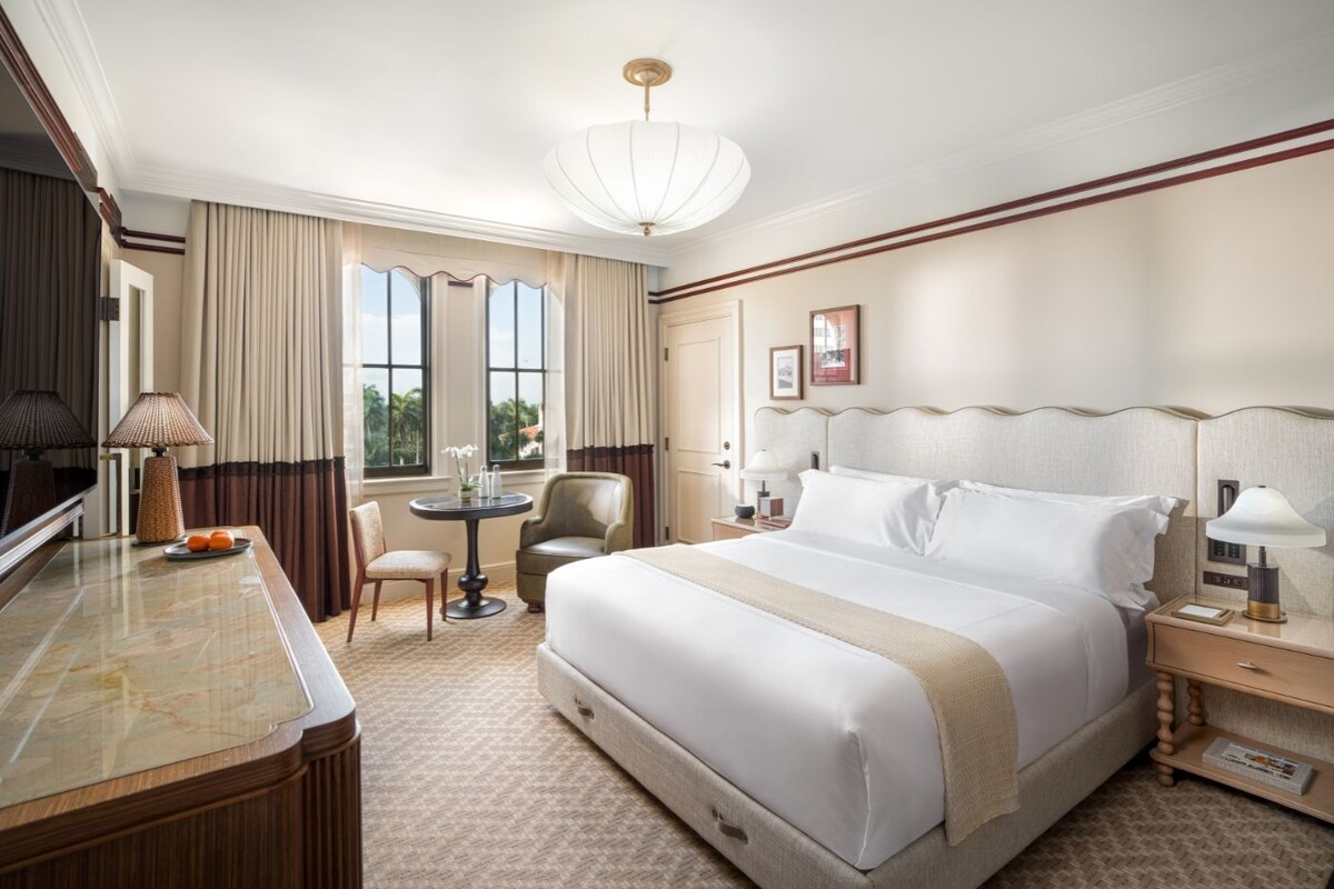 Habitación de hotel con una cama grande, un escritorio con lámpara, una silla, una mesita y una ventana con cortinas.