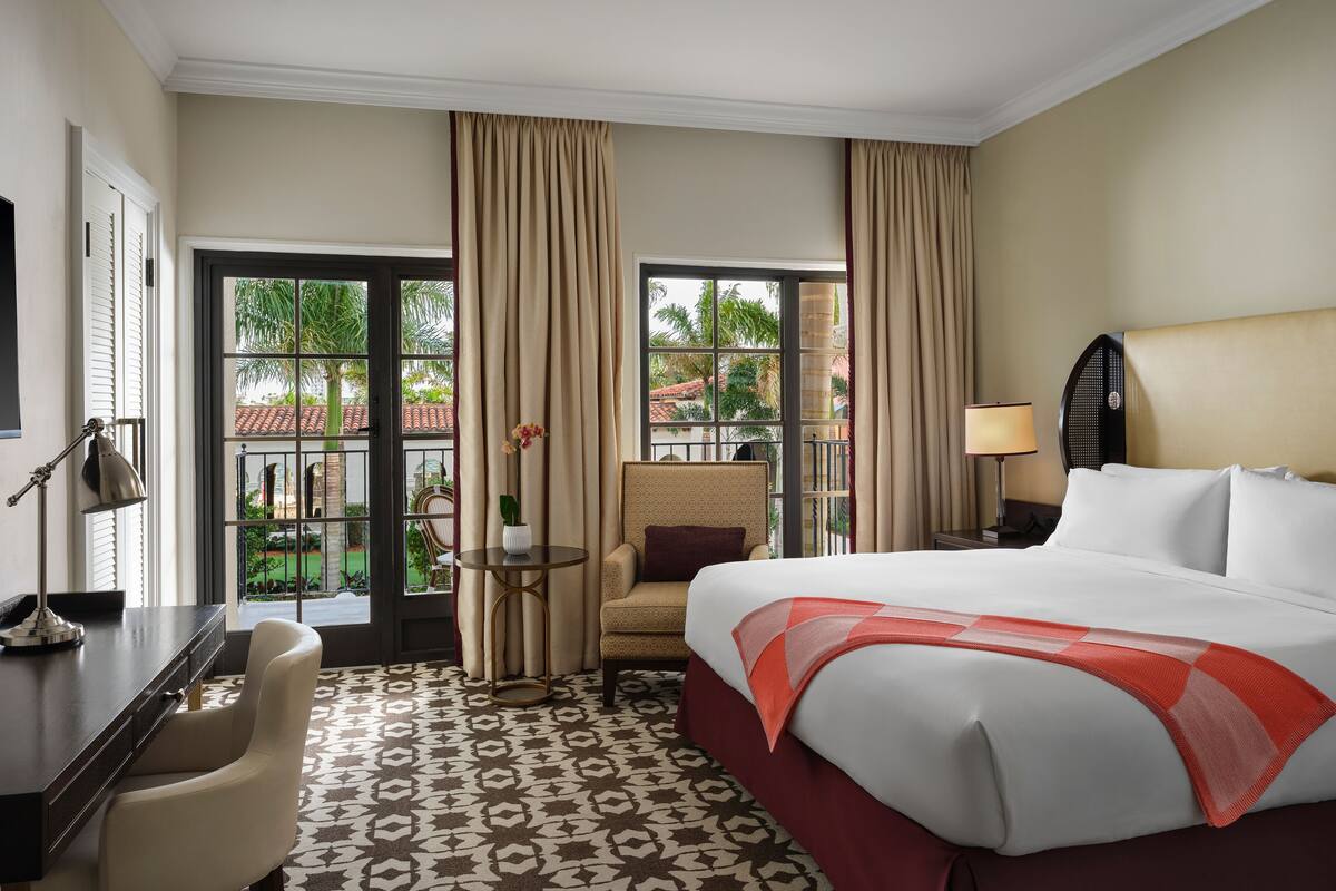 A hotel room with a large bed, a desk, a chair, a small table with a vase, and a view of the outdoors through large windows.