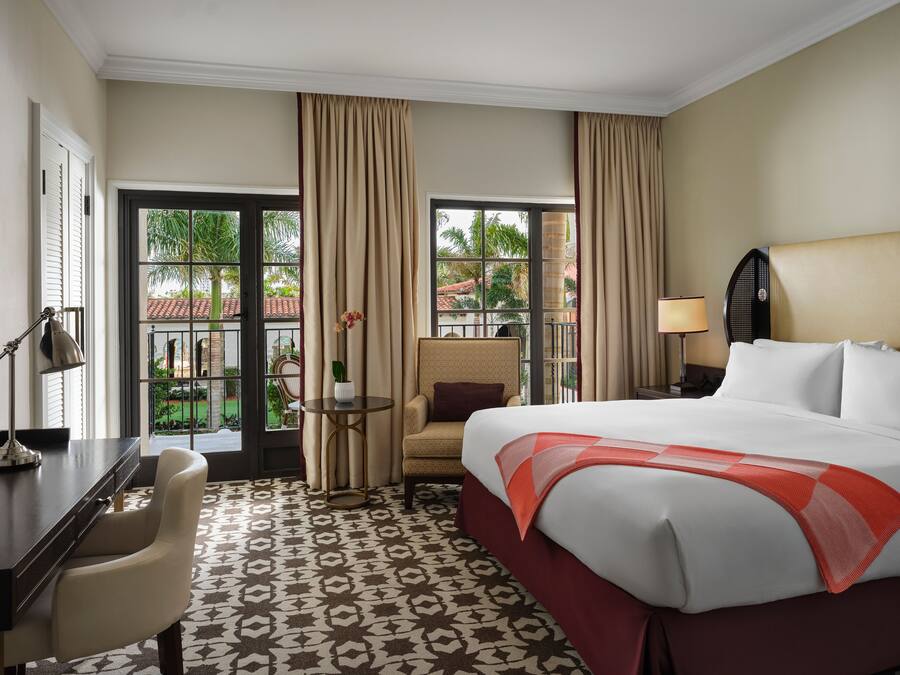 Quarto de hotel com uma cama grande, uma escrivaninha, uma cadeira, uma mesa pequena com um vaso e vista para o exterior através de amplas janelas.