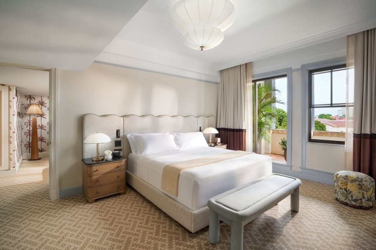 Habitación de hotel con una cama grande, una mesita de noche con lámpara, un banco y vistas al exterior a través de una ventana.