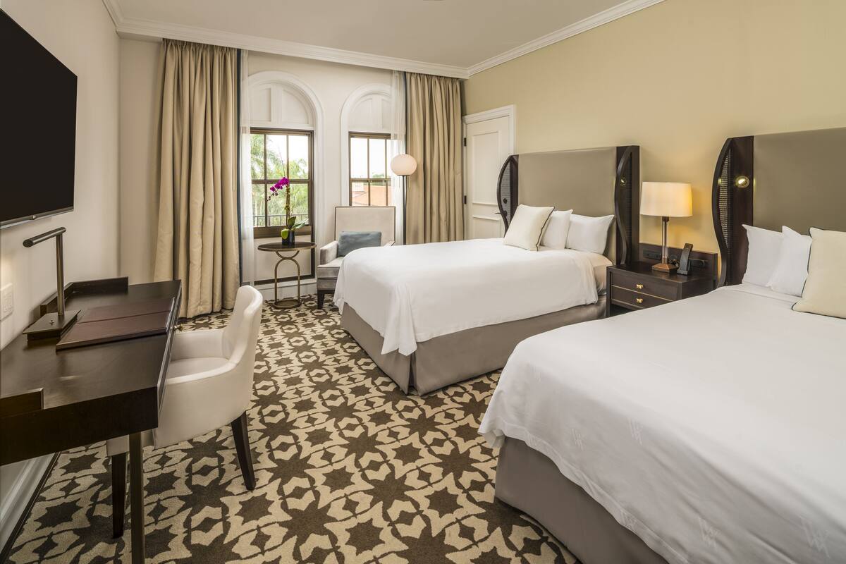 A hotel room with two beds, a desk, a chair, a TV, and a window with curtains.