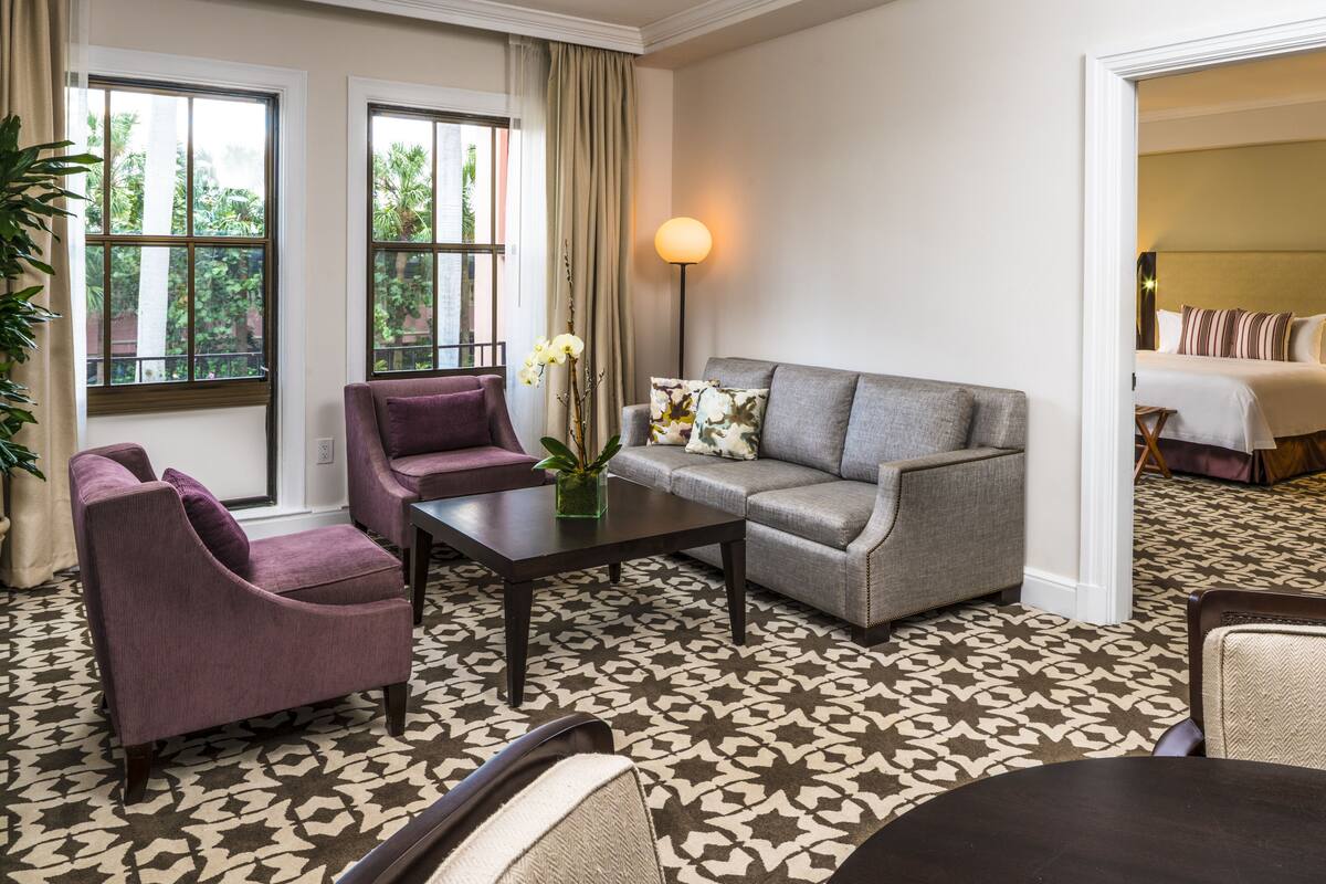 A hotel room with a sofa, armchair, and a small table with a vase of flowers.