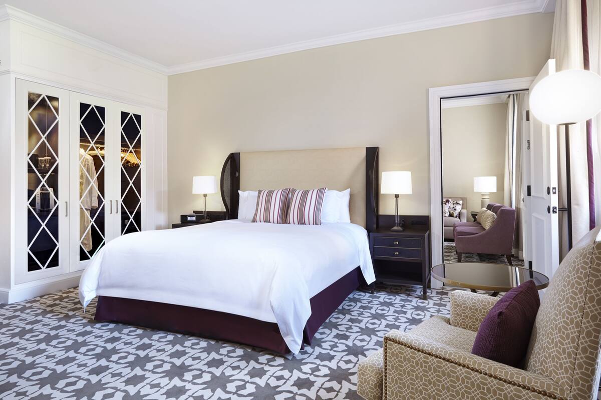 A hotel room with a large bed, a sitting area with a sofa and armchair, a glass coffee table, a wardrobe with mirrored doors, and a large mirror.