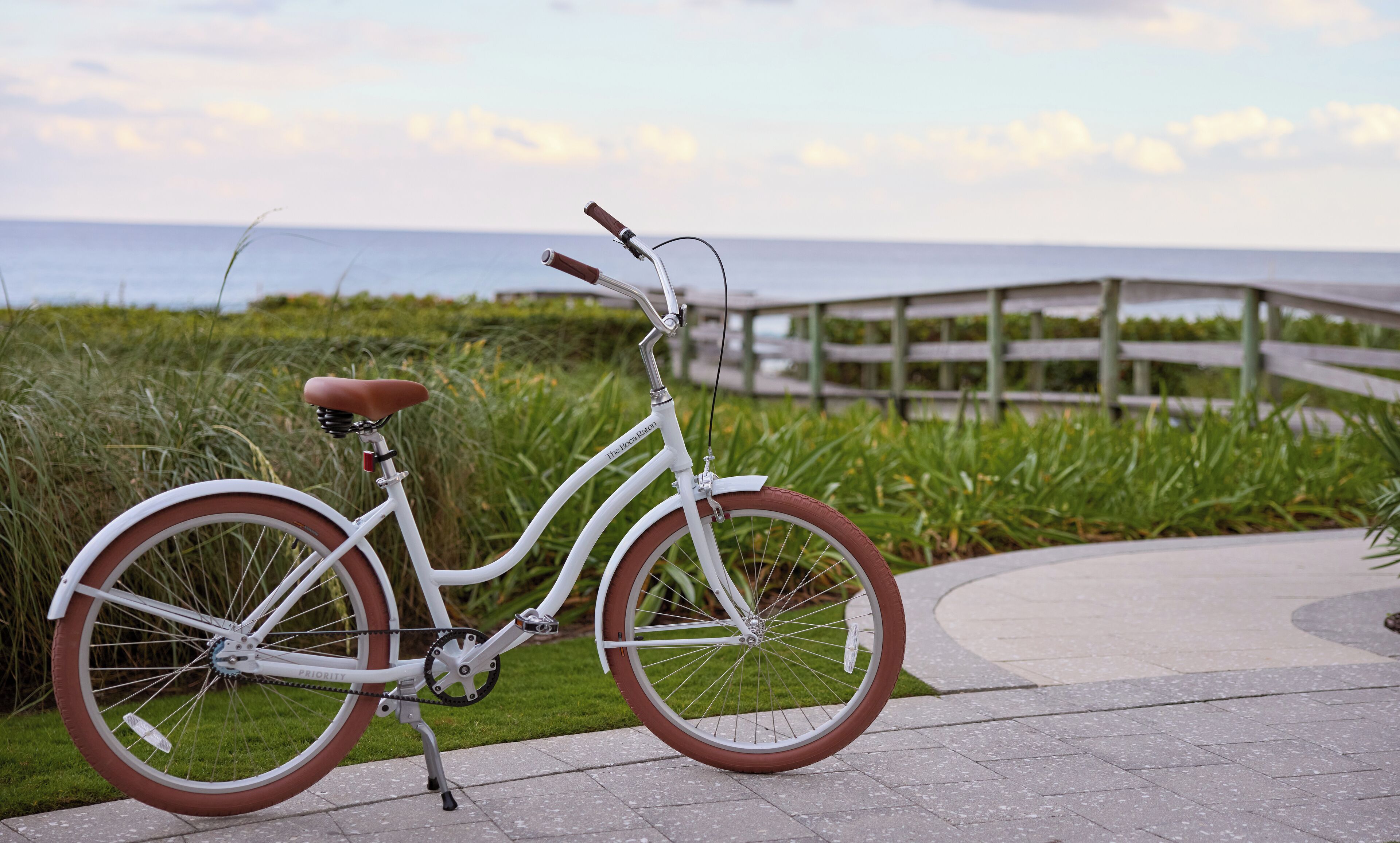 A white bicycle with a brown seat and handlebars, parked on a paved path with a wooden bridge and grassy area in the background.