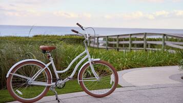 A white bicycle with a brown seat and handlebars, parked on a paved path with a wooden bridge and grassy area in the background.