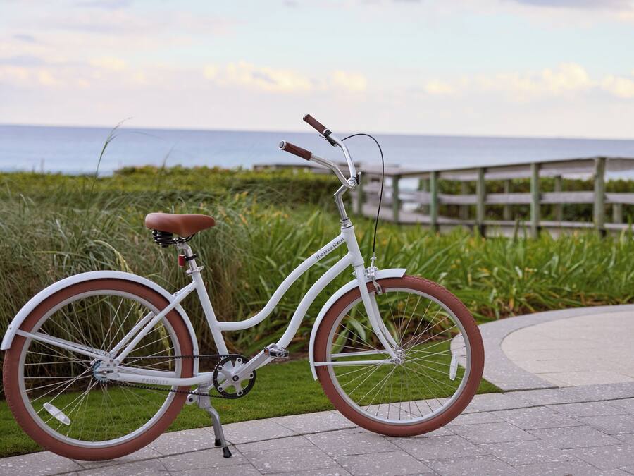 A white bicycle with a brown seat and handlebars, parked on a paved path with a wooden bridge and grassy area in the background.