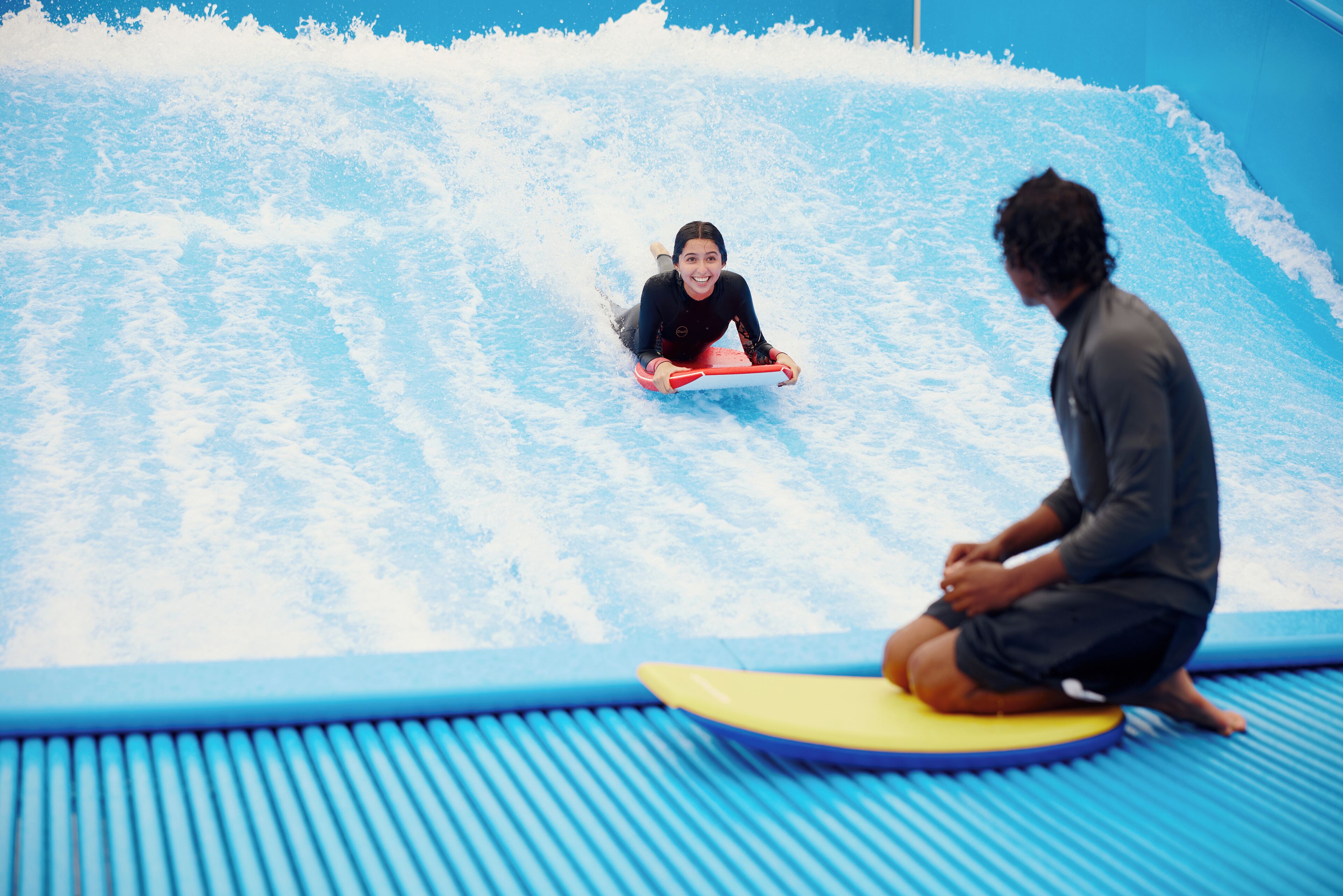 A person sitting on a surfboard in a wave pool, another person standing nearby, and a blue wave pool with a blue floor.