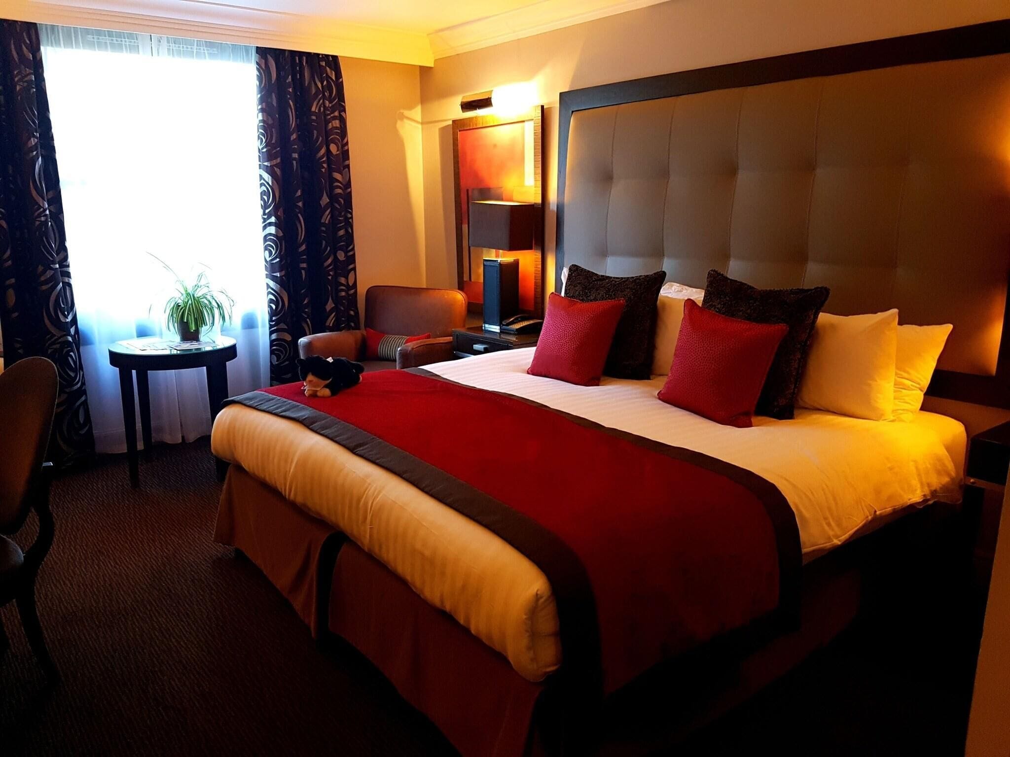 A hotel room with a large bed, a bedside table, a chair, a desk, and a lamp.