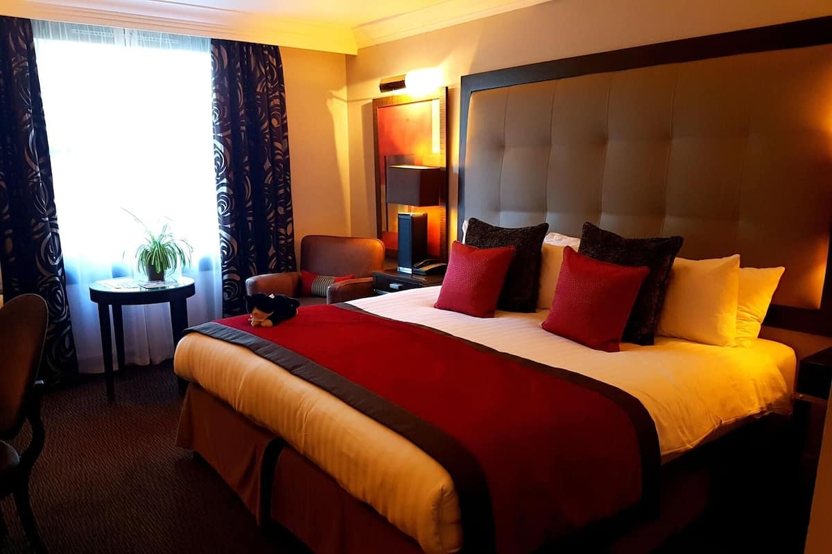A hotel room with a large bed, a bedside table, a chair, a desk, and a lamp.