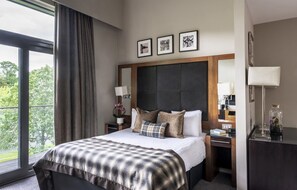 A hotel room with a large bed, a nightstand with a lamp, a desk with a lamp, and a view of trees through the window. - Norton House Hotel & Spa (Newbridge)