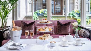 A three-tiered dessert stand with pastries and sandwiches, a champagne bucket, and tea service on a table in a room with a sofa and a view of a garden through large windows.