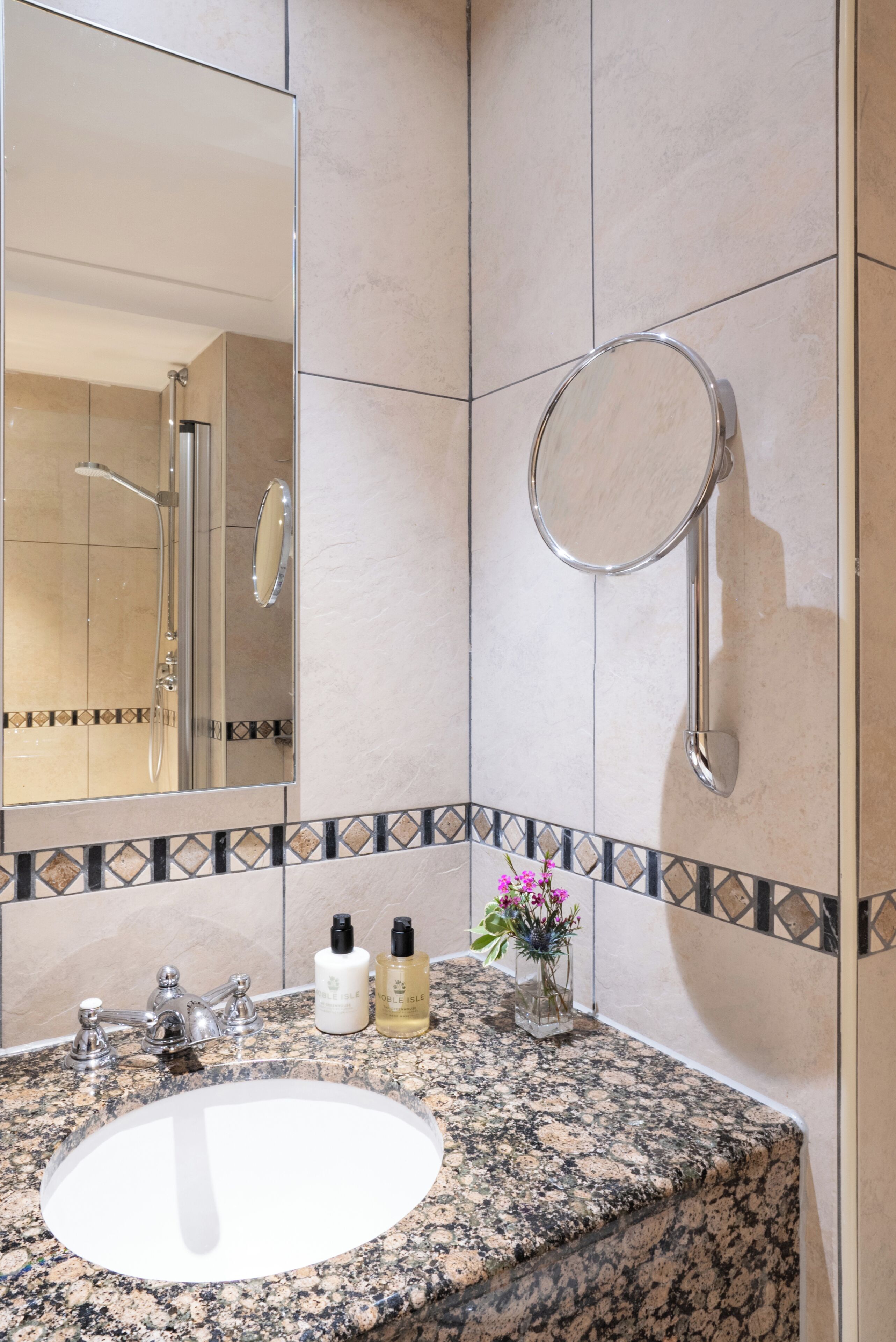 Een badkamer met een wastafel met een granieten blad, een spiegel, een handdouche en een kleine vaas met bloemen.