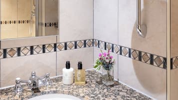 Een badkamer met een wastafel met een granieten blad, een spiegel, een handdouche en een kleine vaas met bloemen.