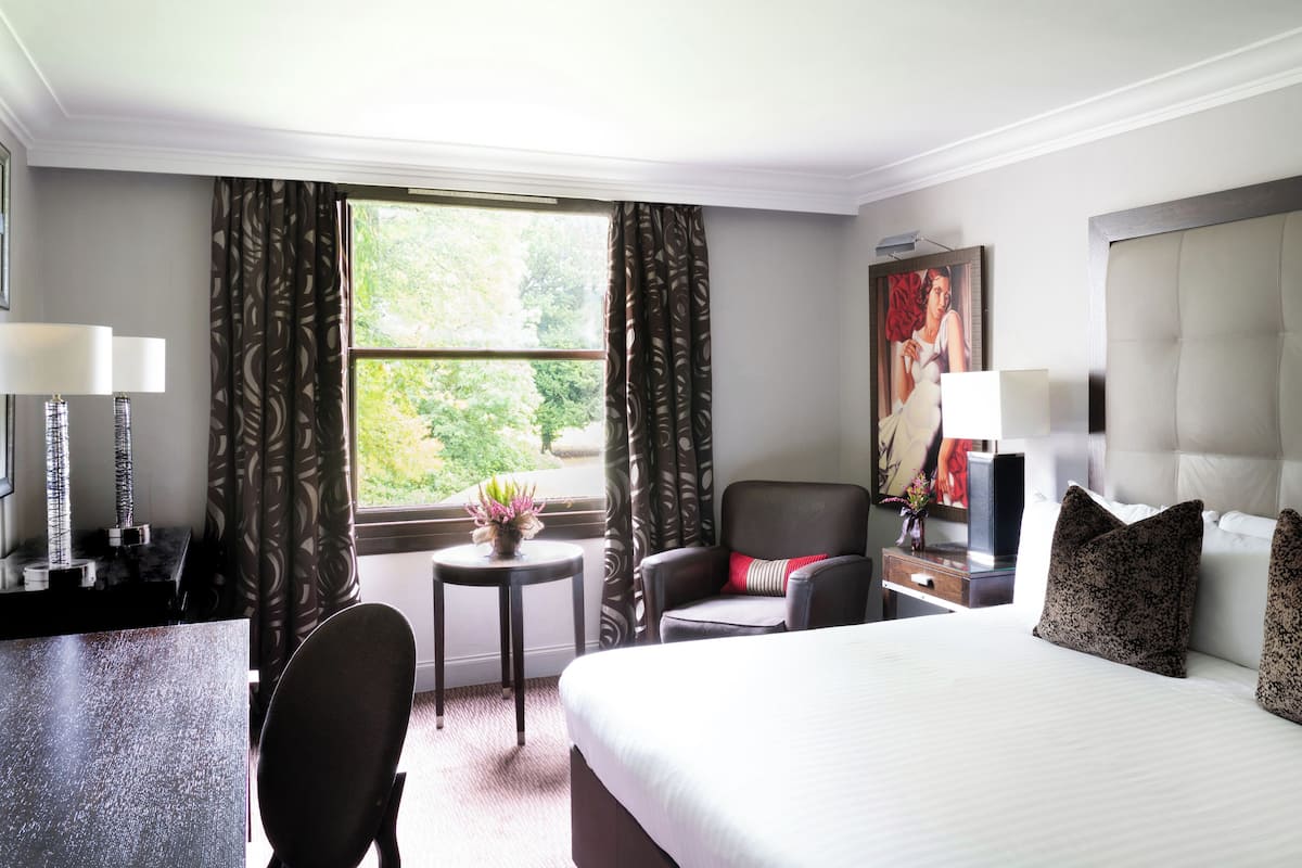A hotel room with a bed, a desk, a chair, a small table with a vase, and a window with curtains.