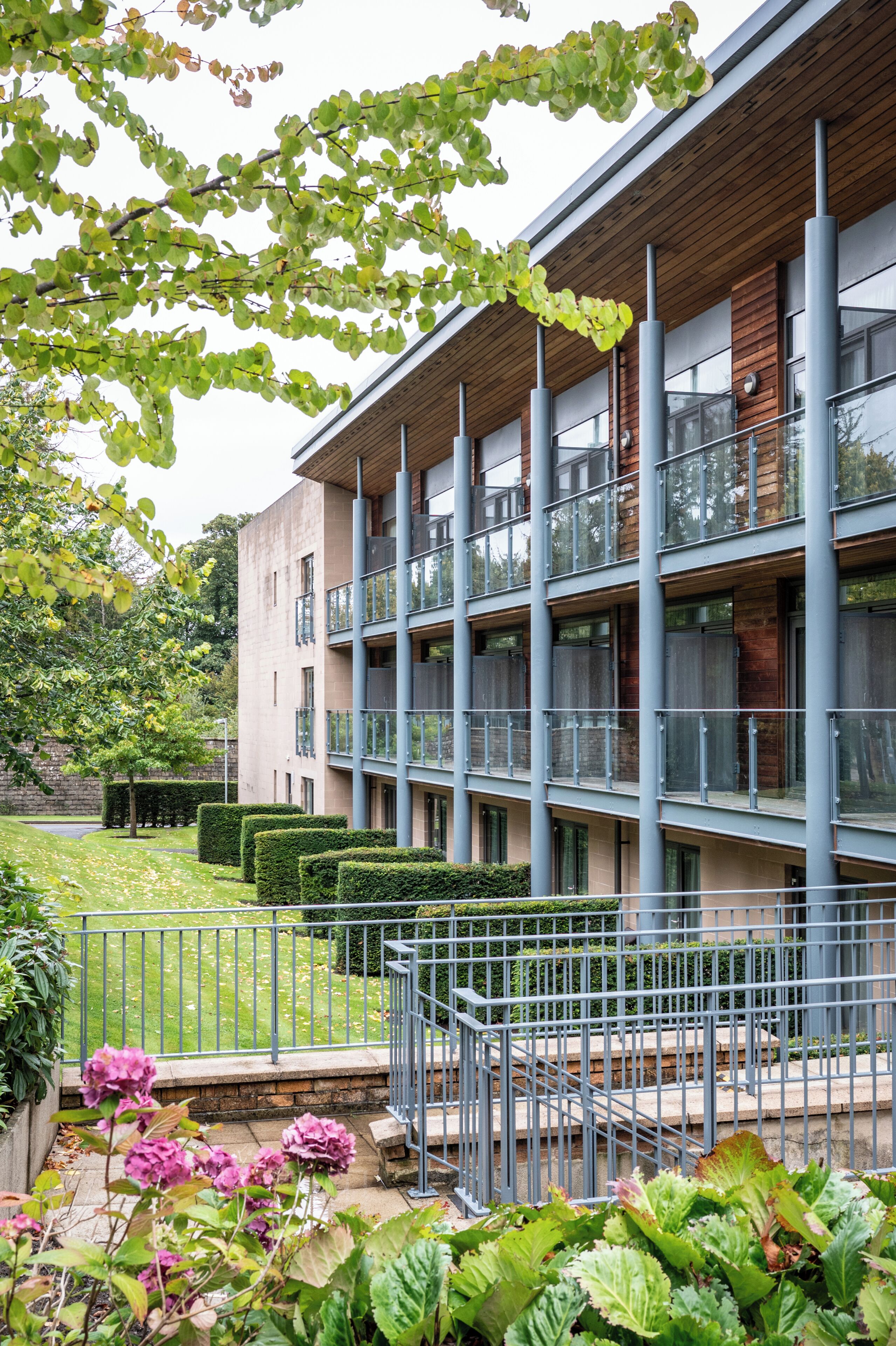 A multi-story building with balconies, a courtyard with hedges, and a staircase leading to the entrance.