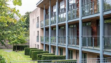 A multi-story building with balconies, a courtyard with hedges, and a staircase leading to the entrance.