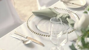 A table set with a clear glass, a silver spoon, a fork, and a knife, with a green napkin and a beaded necklace on the table.