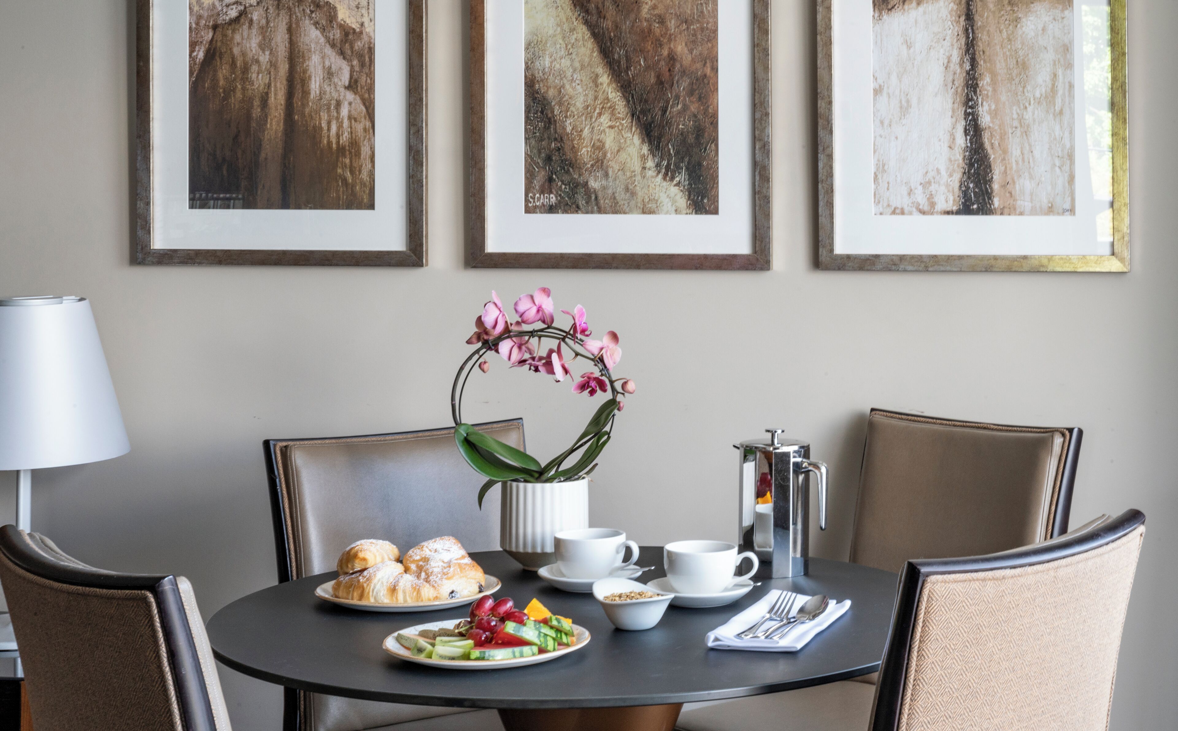 A dining table with croissants, fruit, and coffee cups, surrounded by chairs, with framed artwork on the wall.
