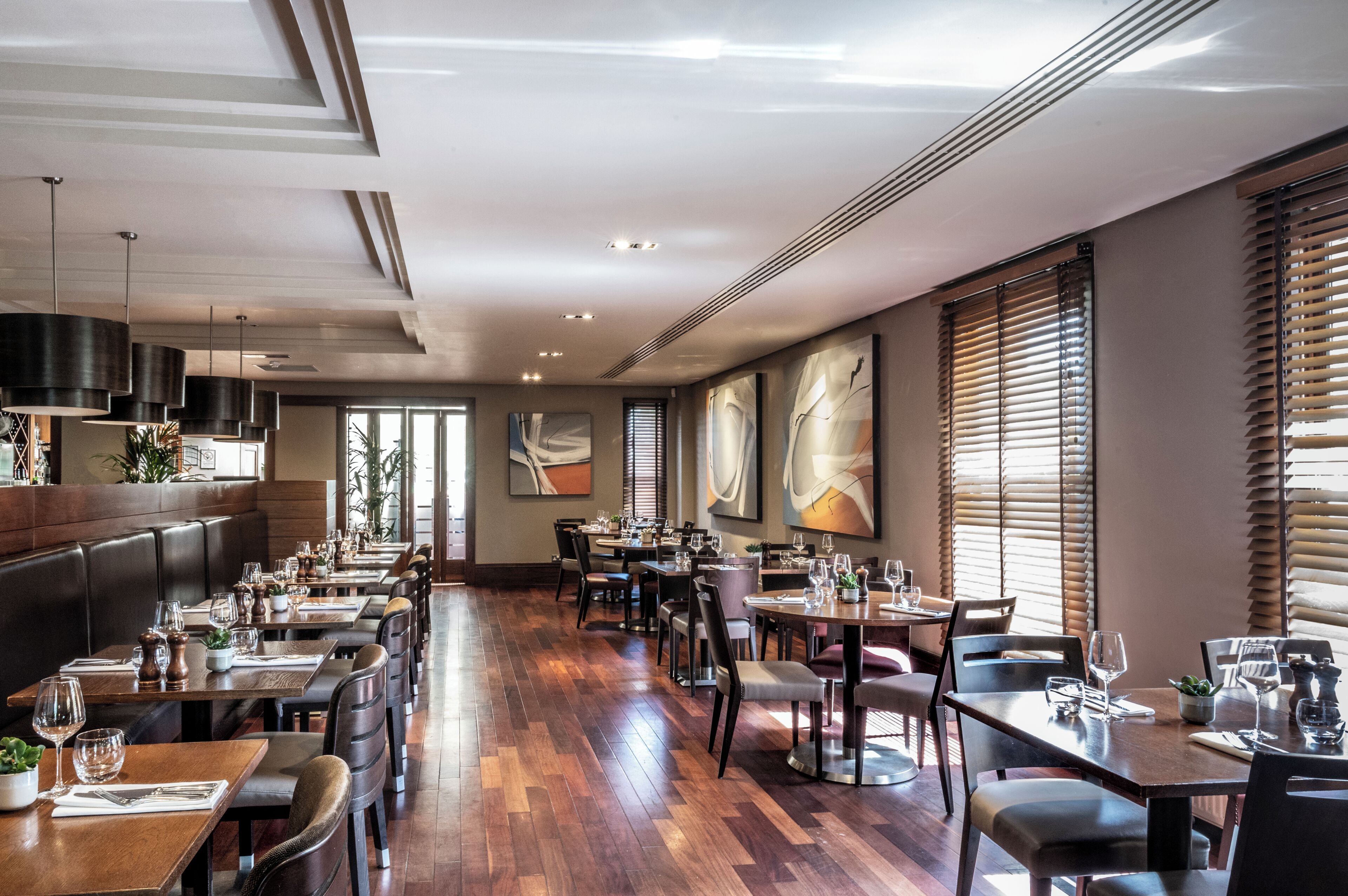 A restaurant with wooden flooring, multiple tables and chairs, and large windows with blinds.
