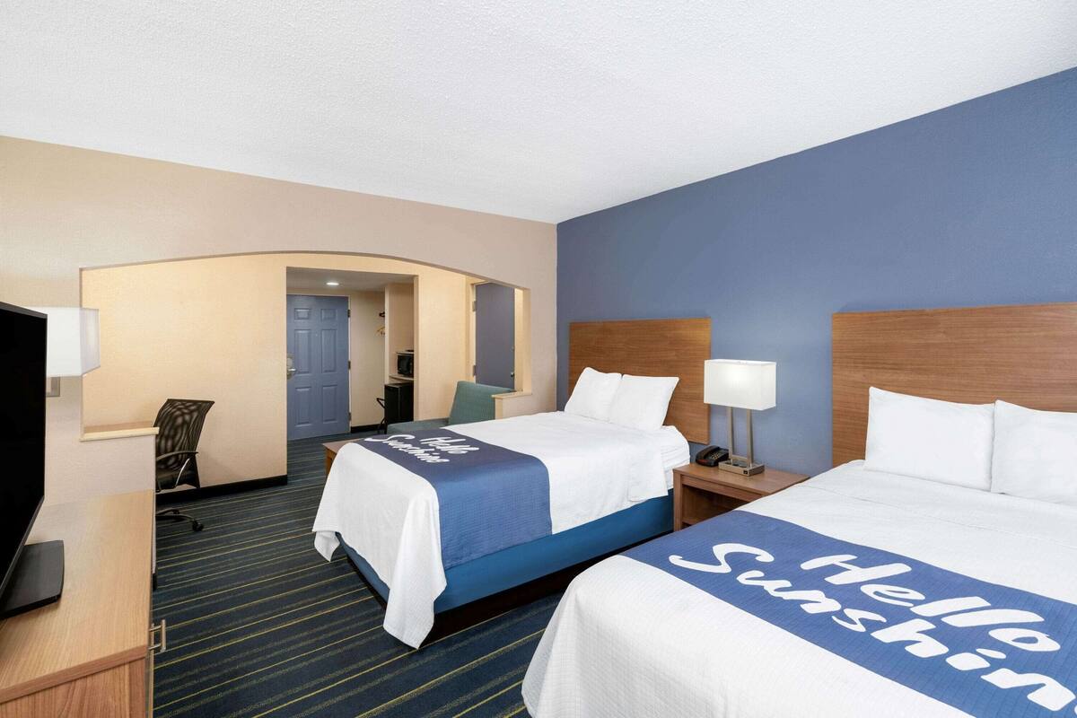 A hotel room with two beds, a desk, a TV, and a bathroom visible through an archway.