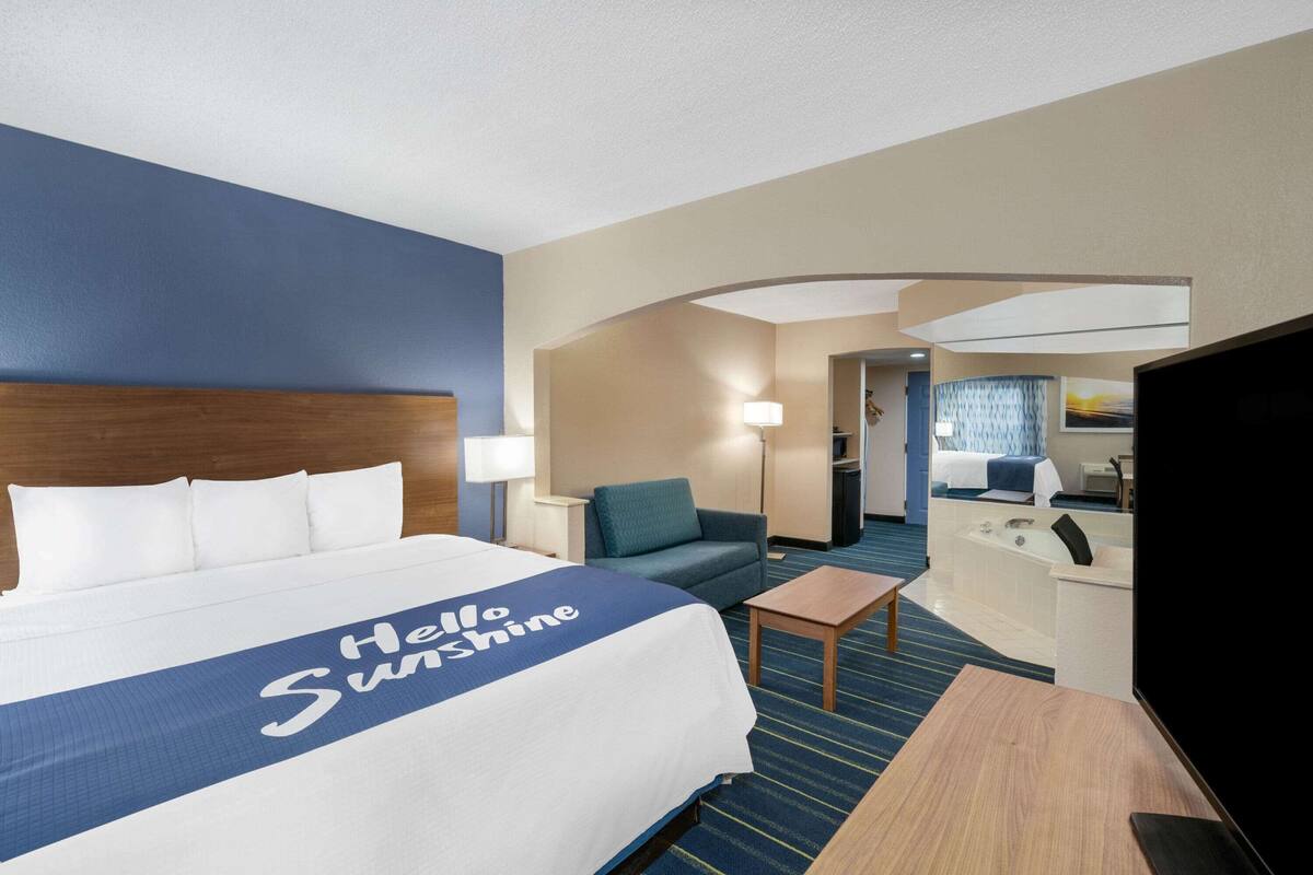 A hotel room with a large bed, a desk, a chair, a TV, and a bathroom visible through an archway.