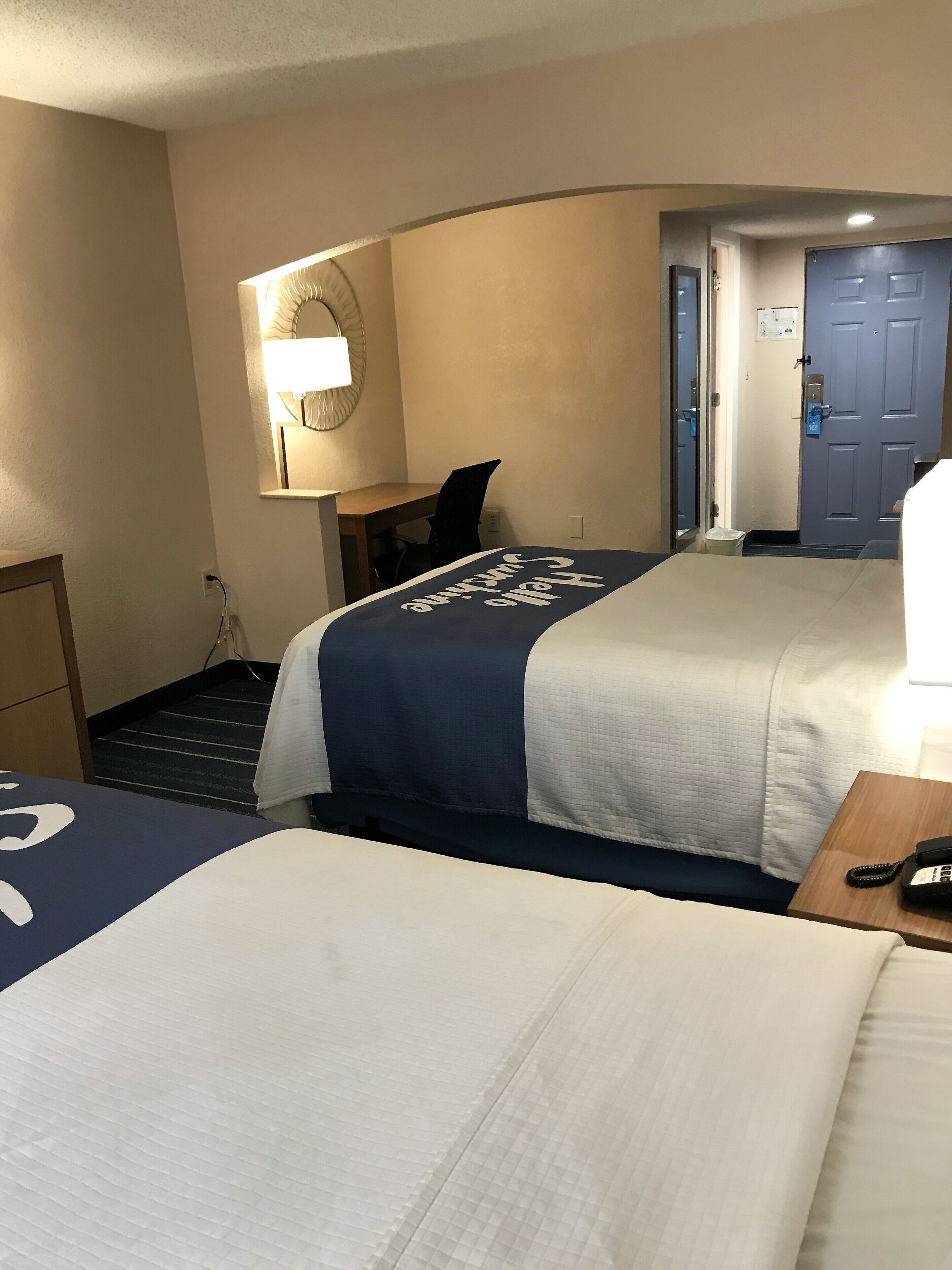 A hotel room with two beds, a desk, a chair, a lamp, and a door.