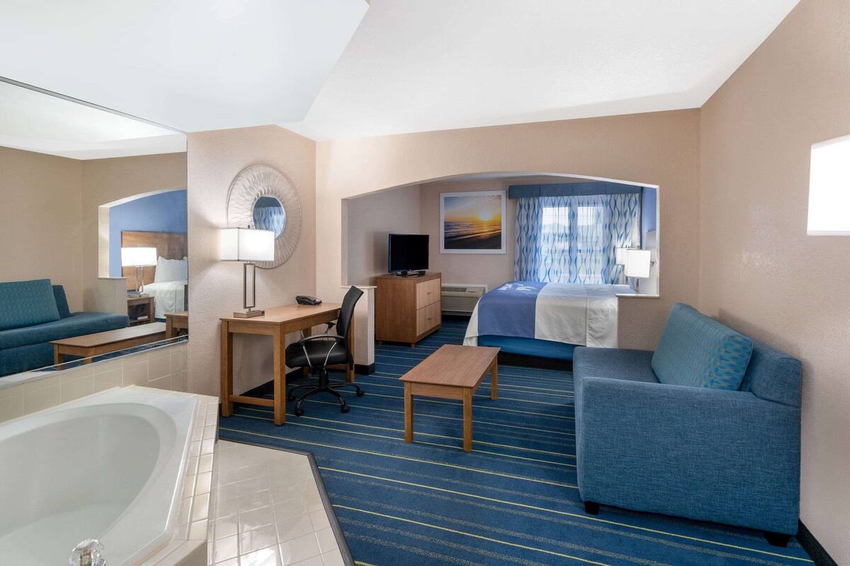 A hotel room with a large bed, a sitting area with a sofa and armchair, a desk with a chair, a TV, and a bathroom with a bathtub.