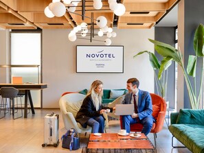 Eine moderne Hotelhalle mit Holzbalkendecke, einem Novotel-Schild, einem grünen Sofa, einem Tisch mit einem Laptop und einer Tasse sowie einem Koffer auf dem Boden.