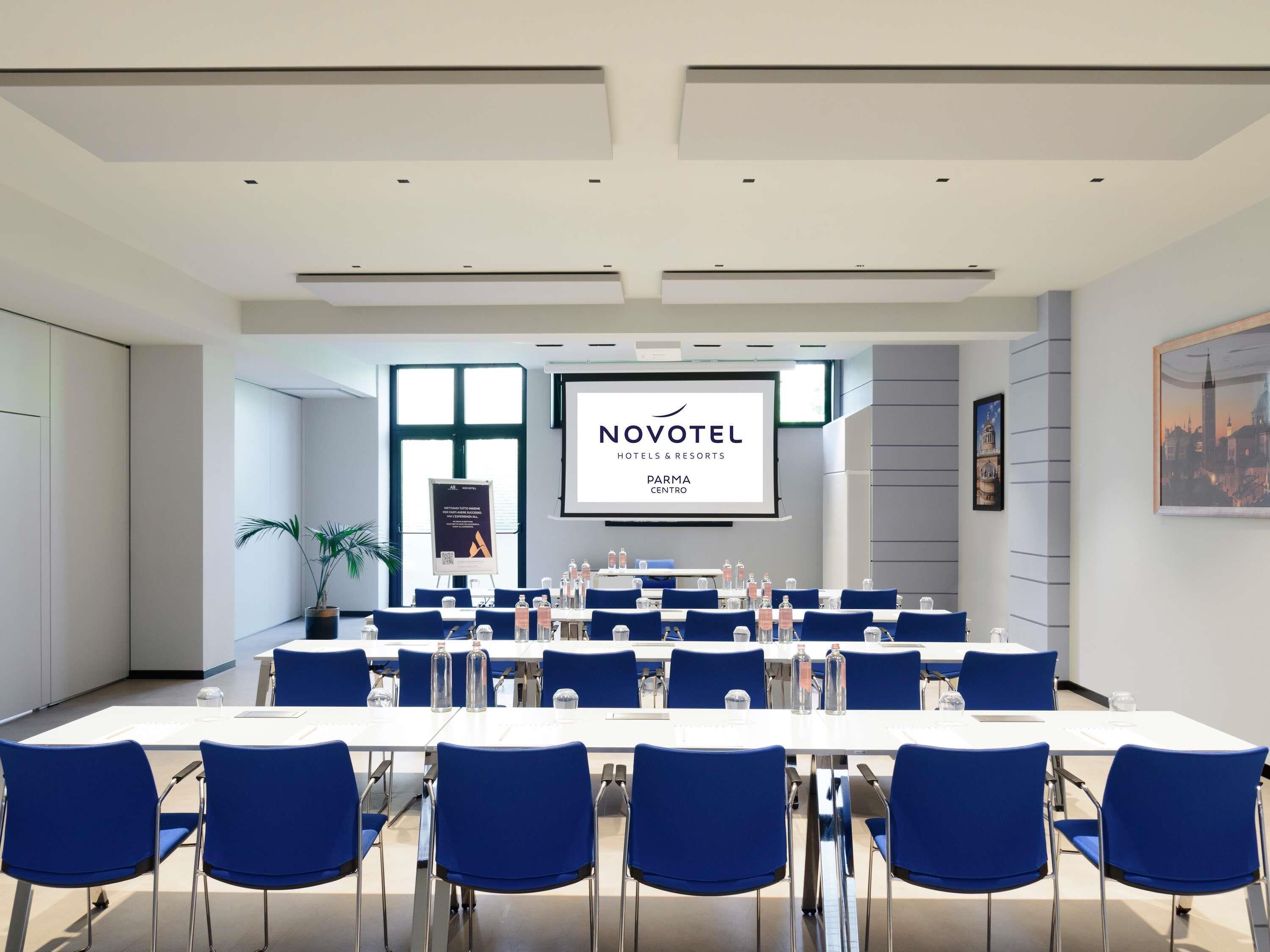 Une salle de conférence dotée d’un grand écran affichant le texte « NOVOTEL Hotels & Resorts Parma Centro » et d’un présentoir.