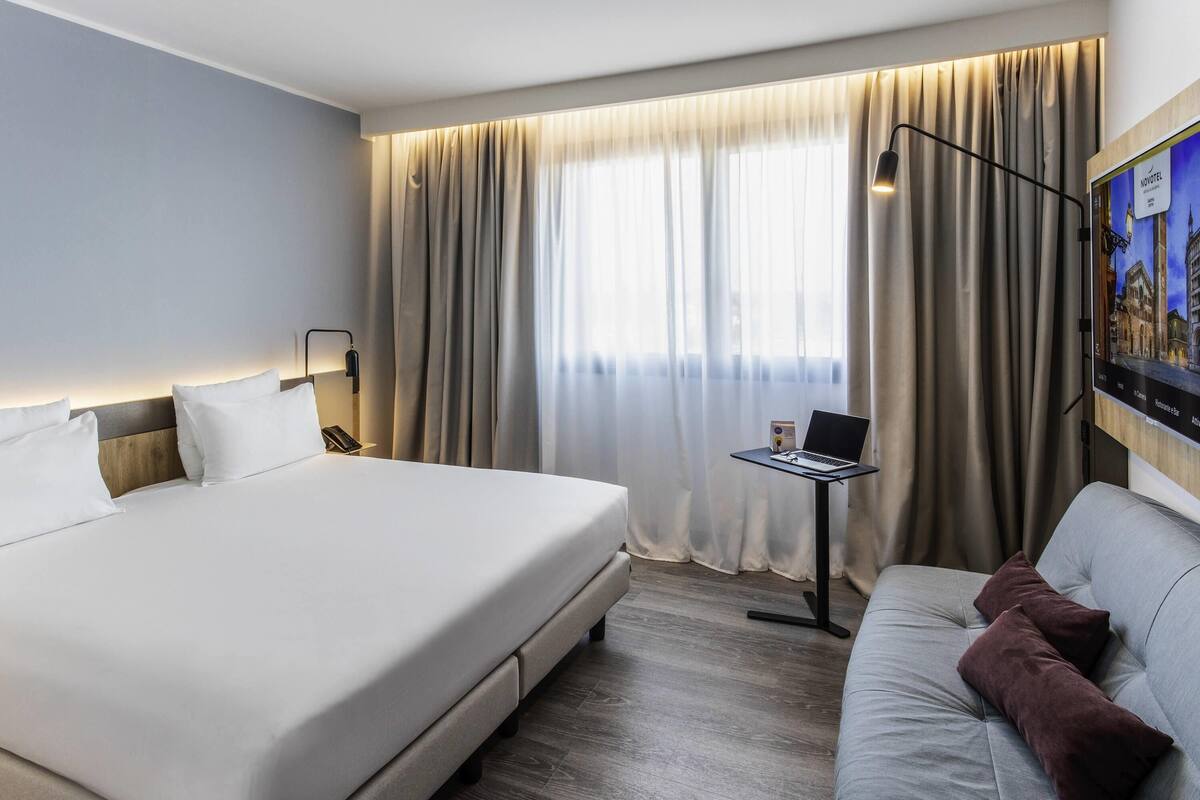A hotel room with a large bed, a desk with a laptop, a sofa, and a large window with curtains.