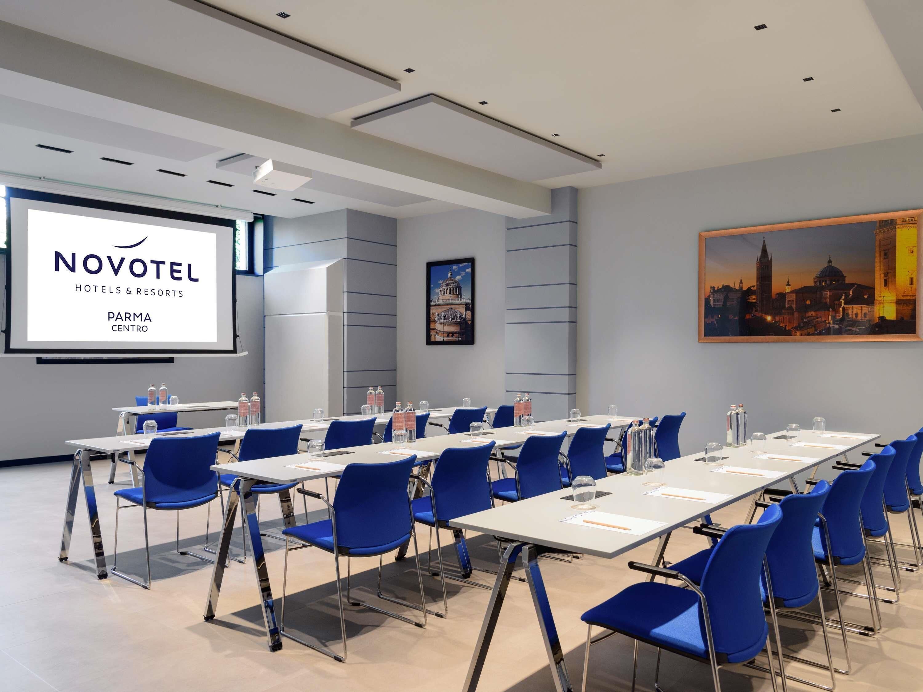 Un salón de conferencias con una pantalla grande mostrando 'NOVOTEL HOTELS & RESORTS PARMA CENTRO', sillas azules, mesas con botellas de agua y un cuadro de un paisaje urbano en la pared.