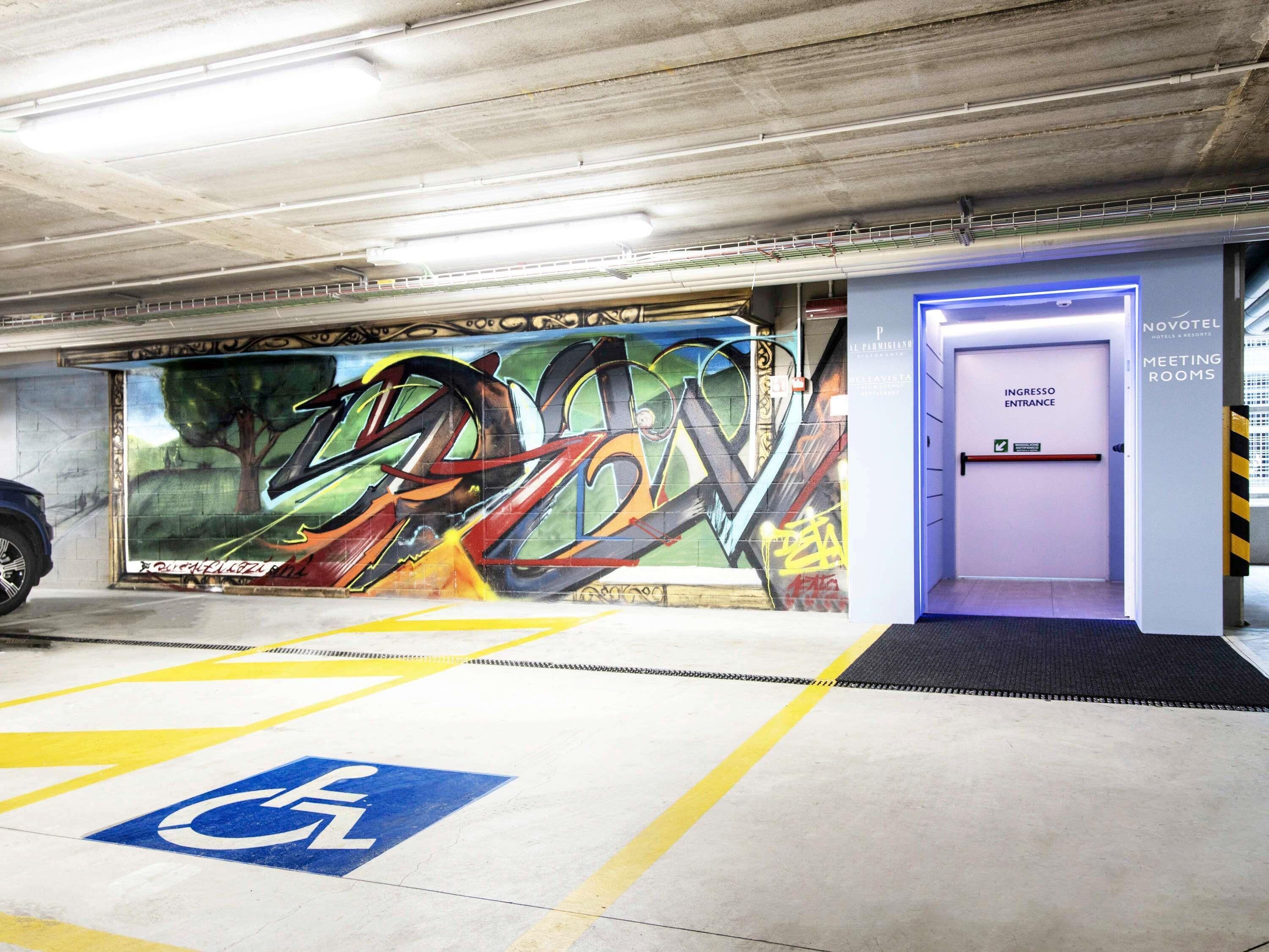 A parking garage with a wheelchair-accessible space, a Novotel hotel entrance, and a graffiti-covered wall.