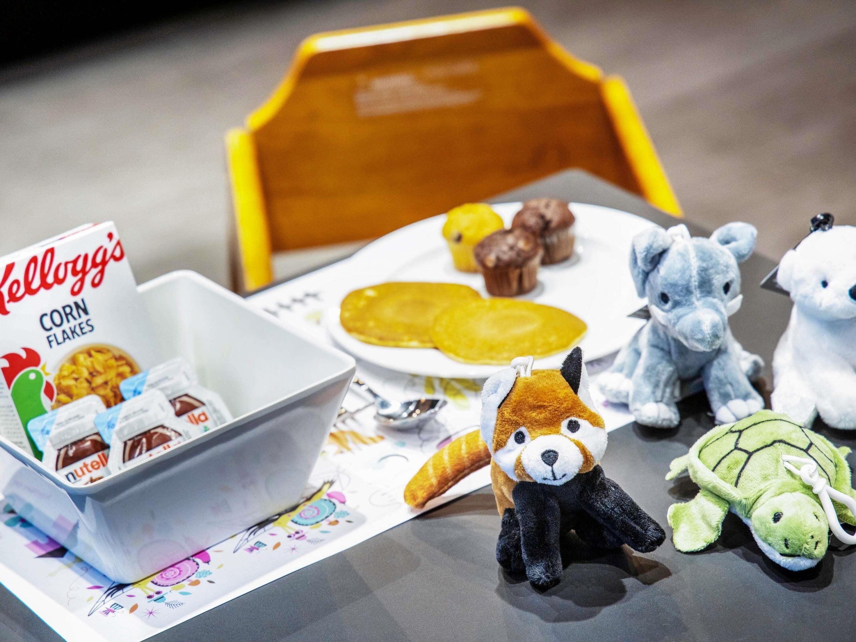 A table with a bowl of cereal, a plate with pancakes, and plush toys.