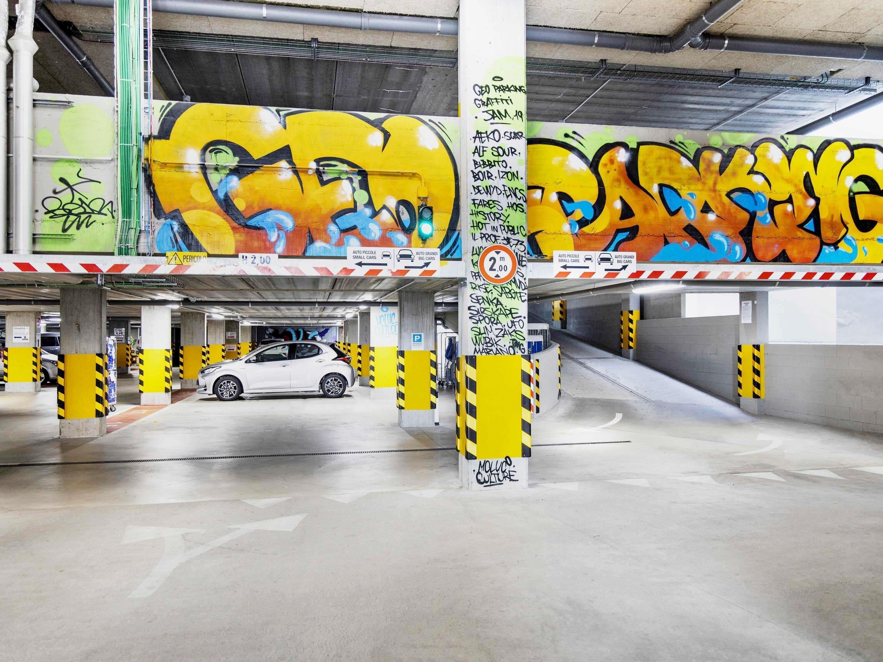 Un parking souterrain dont les murs et les piliers sont couverts de graffitis, une voiture blanche garée au loin, et un panneau indiquant une hauteur maximale de 2,00 mètres.