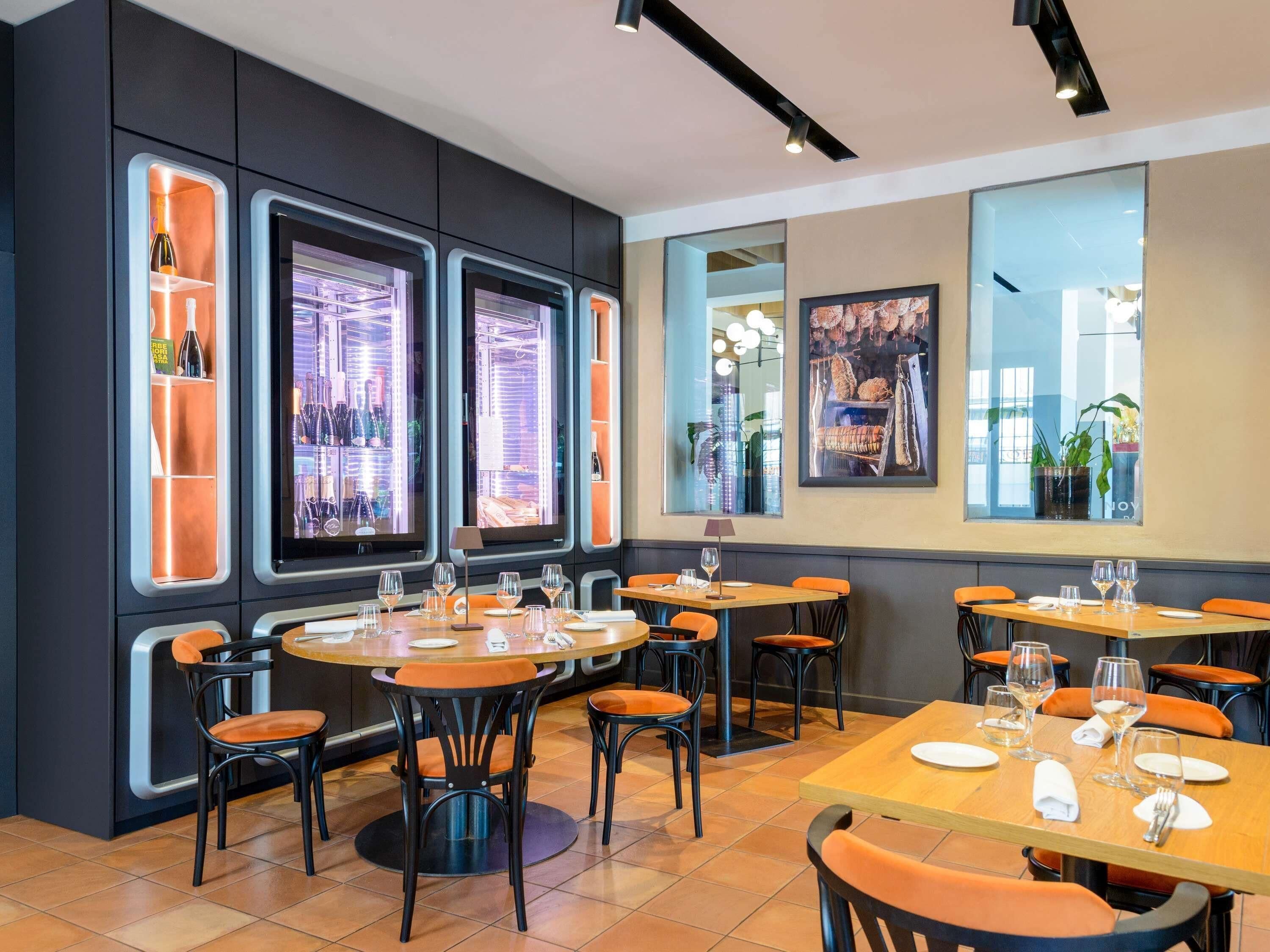 Interno di un ristorante con diversi tavoli e sedie, ampie finestre e scaffali pieni di bottiglie.