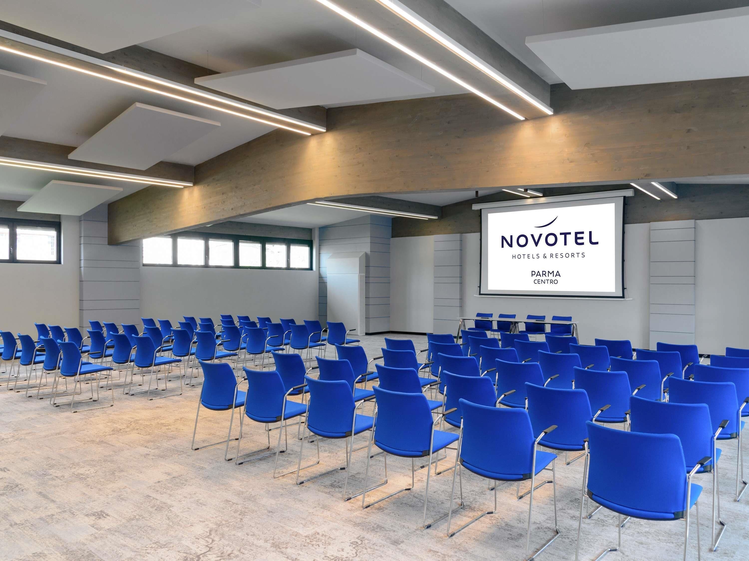 A conference room with rows of blue chairs, a large screen displaying 'NOVOTEL HOTELS & RESORTS PARMA CENTRO', and a modern ceiling design.