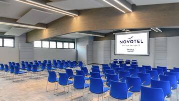 Een vergaderzaal met rijen blauwe stoelen, een groot scherm waarop 'NOVOTEL HOTELS & RESORTS PARMA CENTRO' staat, en een modern plafondontwerp.