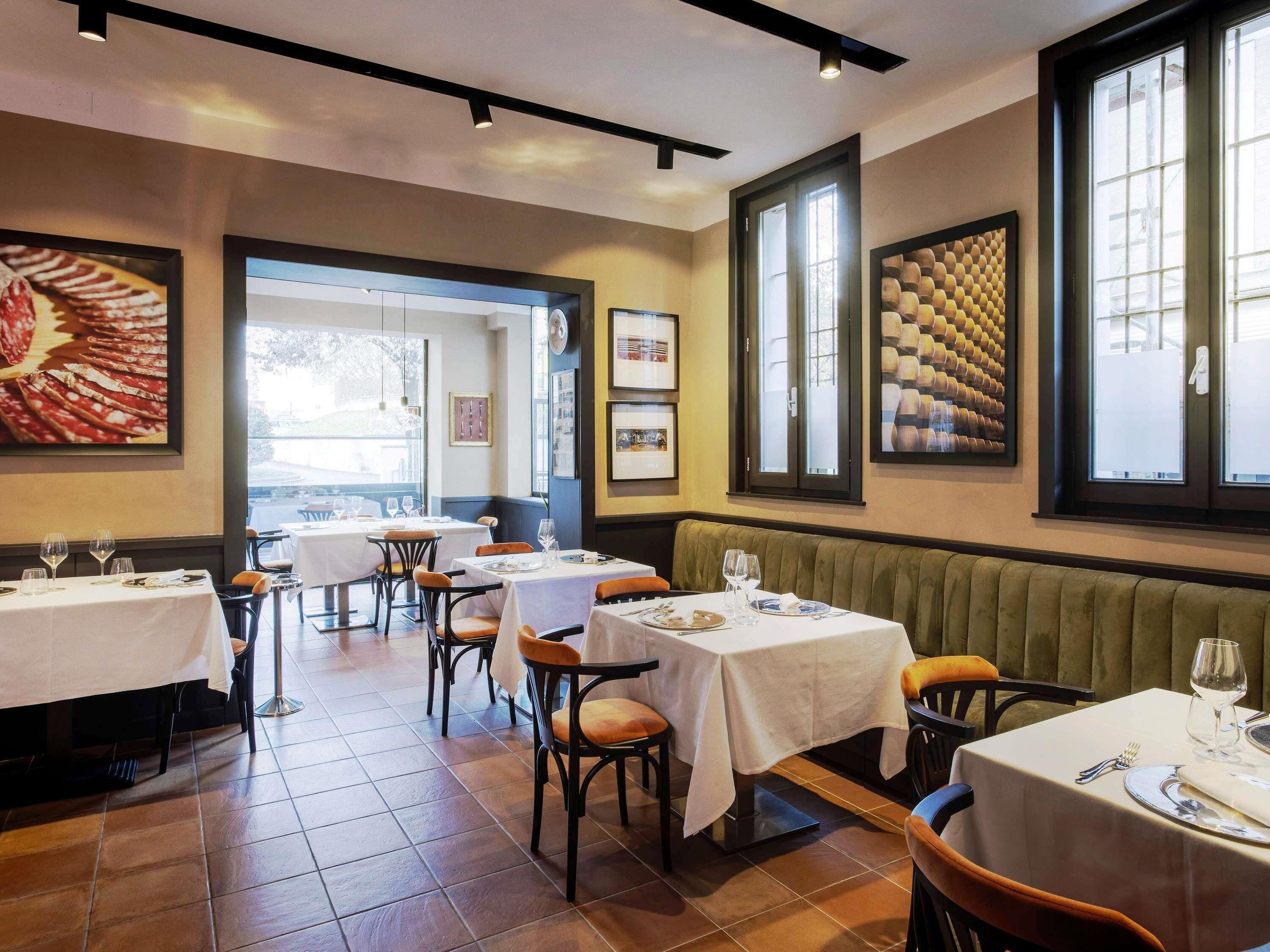 A restaurant interior with tables set for dining, framed artwork on the walls, and large windows allowing natural light.