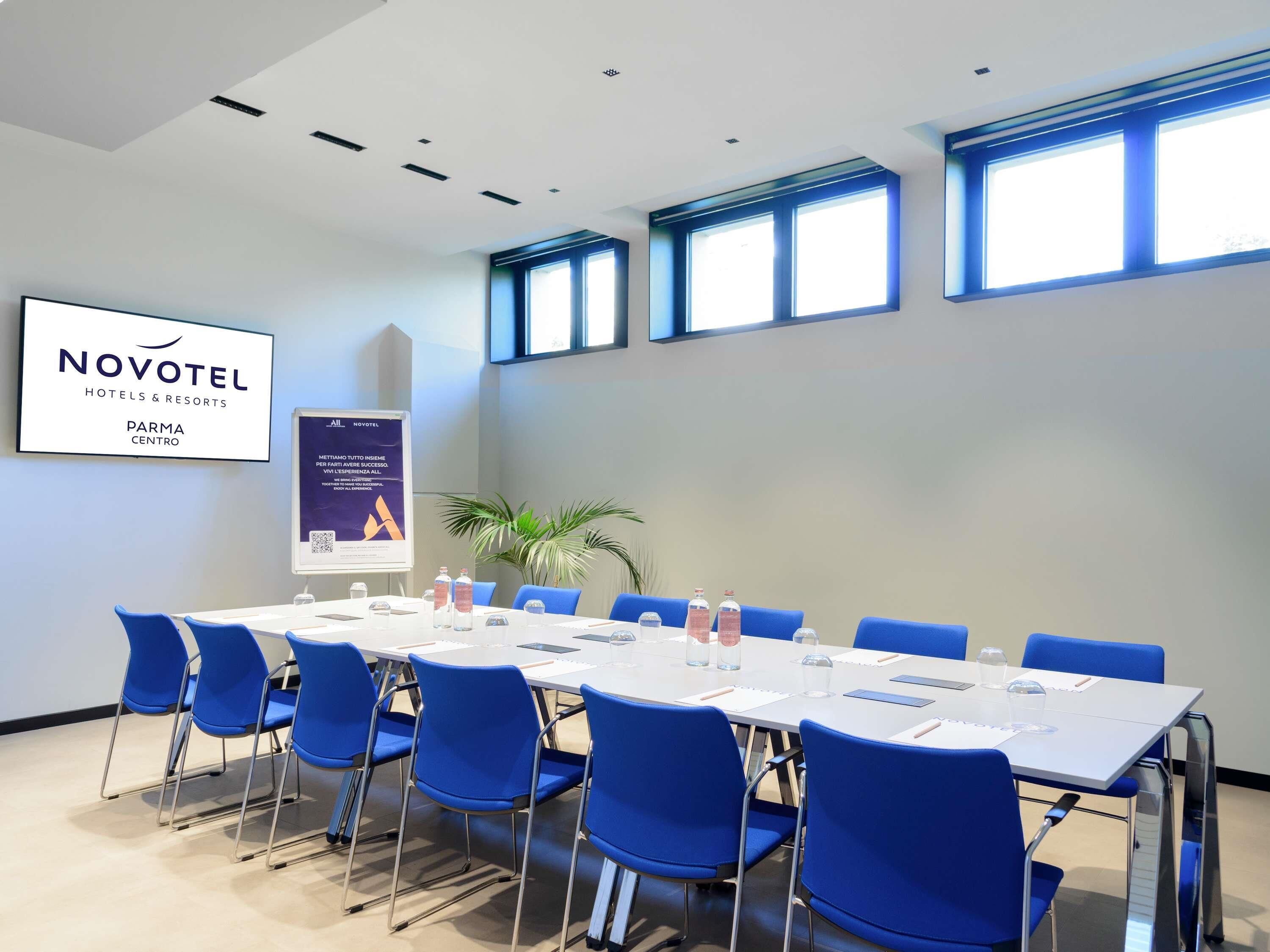 Ein Konferenzraum mit einem langen Tisch, blauen Stühlen und einem Banner für Novotel Hotels & Resorts.