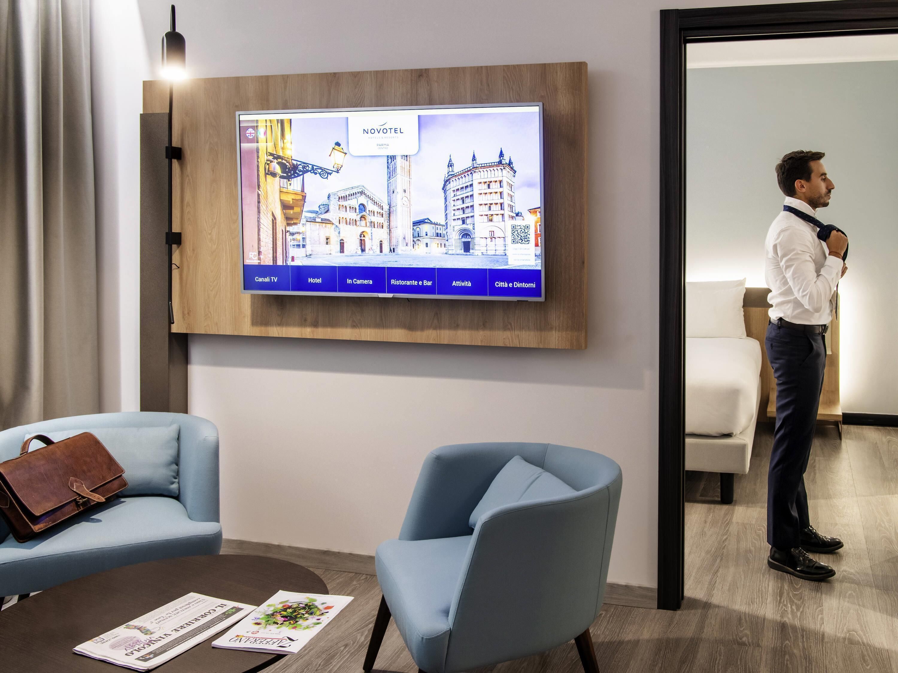 Una camera d'albergo con una TV che mostra una vista sulla città, una poltrona blu, una borsa in pelle marrone e un pavimento in legno.